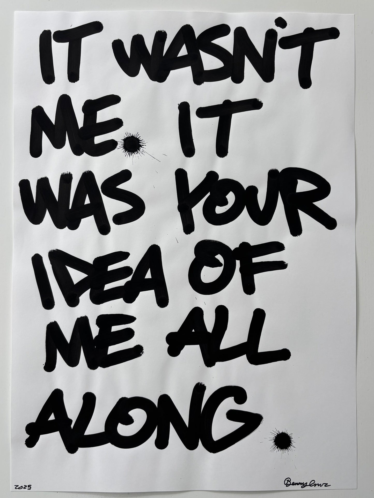 A typographic art print featuring black hand-painted brush lettering on a white background, reading 'IT WASN'T ME, IT WAS YOUR IDEA OF ME ALL ALONG' with small black ink splatters.