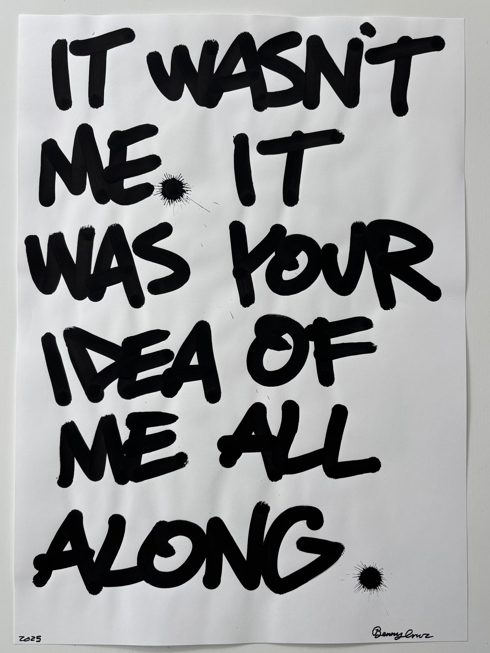 A typographic art print featuring black hand-painted brush lettering on a white background, reading 'IT WASN'T ME, IT WAS YOUR IDEA OF ME ALL ALONG' with small black ink splatters.