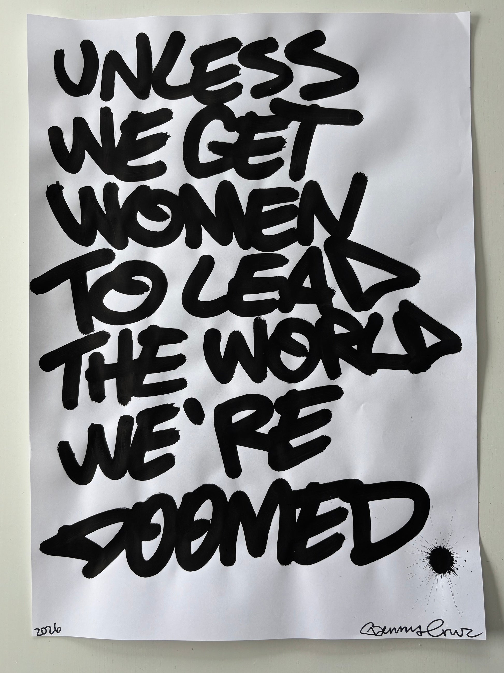 A square white art print or poster featuring bold black graffiti-style lettering that reads 'UNLESS WE GET WOMEN TO LEAD THE WORLD WE'RE DOMED,' photographed against a light neutral background.