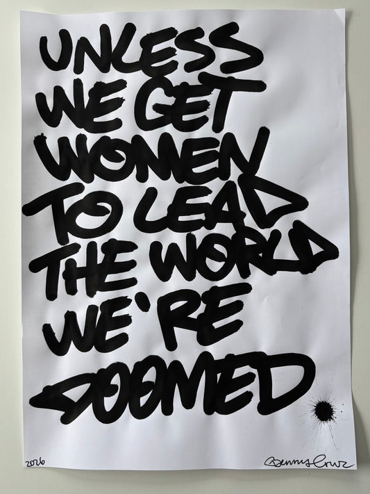 A square white art print or poster featuring bold black graffiti-style lettering that reads 'UNLESS WE GET WOMEN TO LEAD THE WORLD WE'RE DOMED,' photographed against a light neutral background.